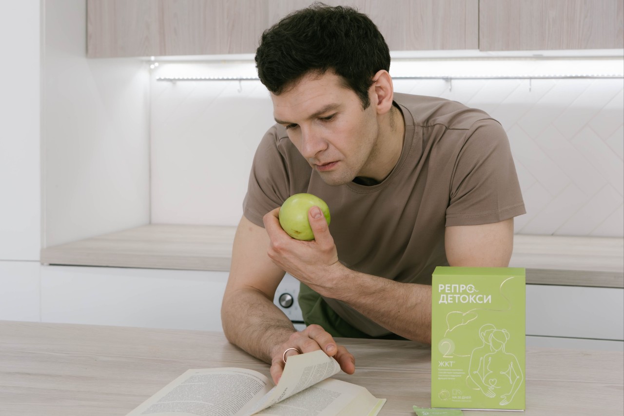 Мужчина с зеленым яблоком и книгой, рядом упаковка продукта для подготовки к зачатию.