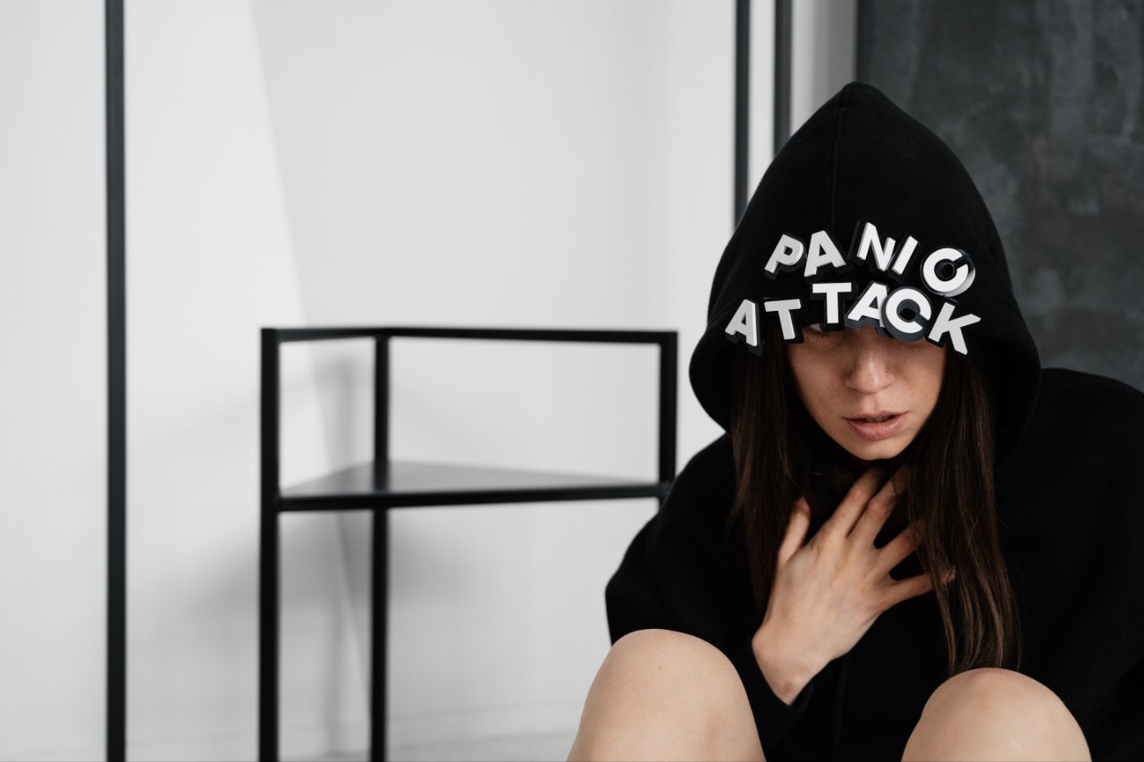Женщина в черном худи с надписью "PANIC ATTACK" выглядит тревожно, сидя на полу.